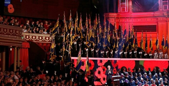A veteran speaks at a podium inside a large hall filled with people. Numerous flags are displayed on the stage, and a poppy motif decorates the background, honoring those who have served in the army.
