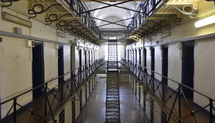 Interior view of a prison cell block with barred doors on both sides and a metal walkway in the center, akin to the disciplined order of the Royal Navy. Overhead lighting casts stark shadows, reminiscent of resettlement facilities designed for structured transition.