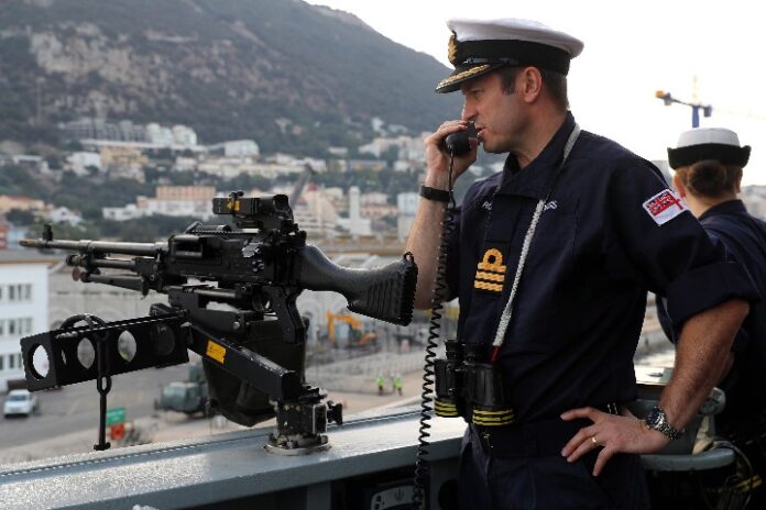 A Royal Navy officer in uniform speaks into a handset beside a mounted machine gun on the ship deck, with a coastal landscape unfolding like a pathfinder's guide in the background.