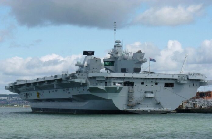 A large, gray Royal Navy aircraft carrier docked at a port under a cloudy sky, showcasing its formidable military presence.