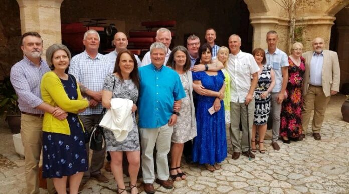 A group of people, including a veteran, stands together outside in a courtyard, posing for a photo. Some are dressed in casual attire while others wear more formal clothing. Stone pavement and arches form the backdrop, reflecting their diverse journeys and stories of resettlement.