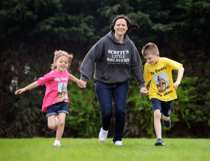 A veteran woman in a gray hoodie is running on grass, holding hands with a girl in a pink shirt and a boy in a yellow shirt. They all appear to be embracing their outdoor adventure with joy.