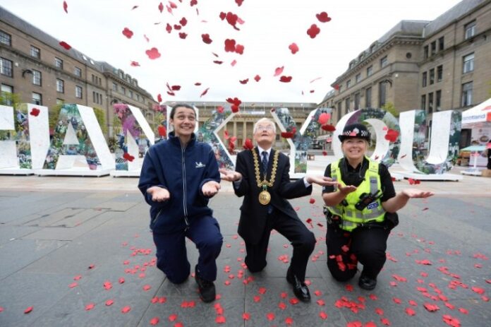 Three people kneel on a city street, surrounded by falling red flower petals, in front of large 