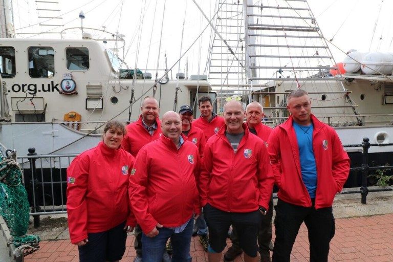 A group of eight people wearing matching red jackets stand in front of a docked military ship.