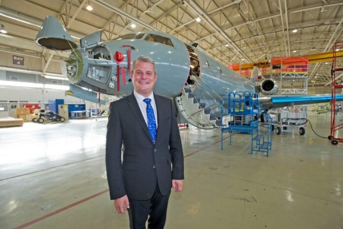 A person in a suit stands in an aircraft assembly hangar, with a partially constructed airplane in the background, highlighting the collaboration between military forces and engineers to innovate aviation solutions.
