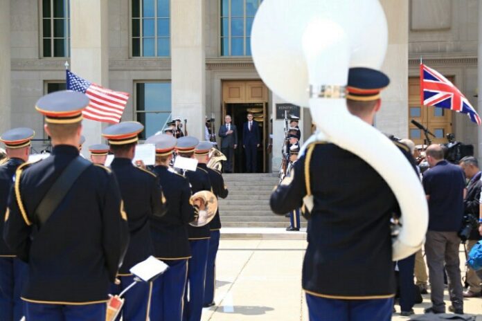 A military band performs outdoors, forming a path for officials at the building entrance. American and British flags flutter alongside veterans' pride, with representatives from both the Army and Royal Navy present.