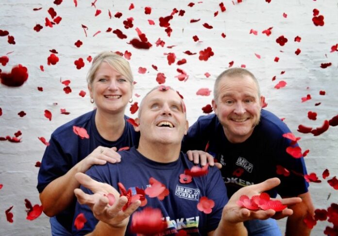 Three veterans wearing blue shirts smile while red petals fall around them against a white brick background, embodying the spirit of camaraderie and resilience.