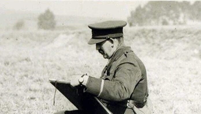 A man in a military uniform sits outdoors, sketching on a large notepad, his focus akin to a pathfinder mapping uncharted territories.