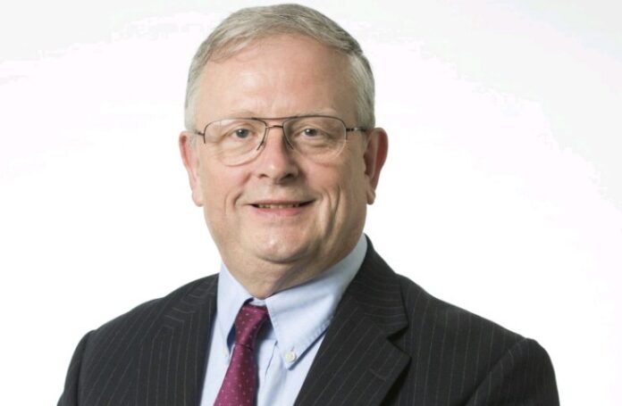 A man wearing glasses, a suit, and a red tie stands against a plain white background, exuding the proud demeanor of an Army veteran.