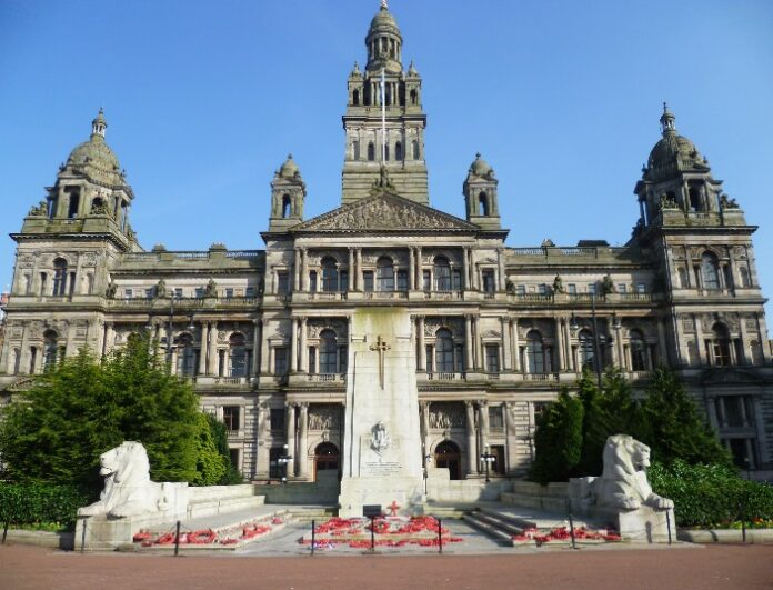Glasgow City Chambers with a World War memorial in the foreground, featuring sculptures and red wreaths honoring the valor of the Army under a clear blue sky.