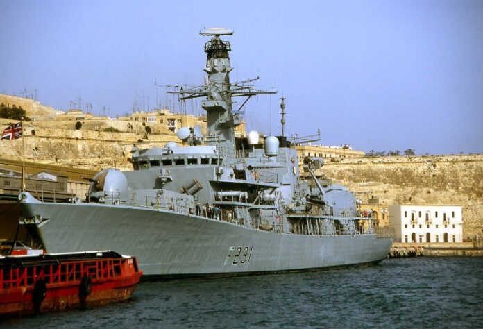 A massive gray military ship docked at the harbor, standing as a steadfast pathfinder against the backdrop of urban buildings, with a small boat quietly drifting nearby.