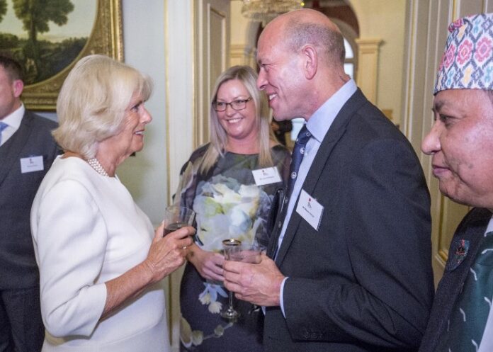 A group of people in formal attire, including a retired Royal Navy veteran, converse at an indoor event. They hold drinks and wear name tags as they share stories and enjoy the evening.