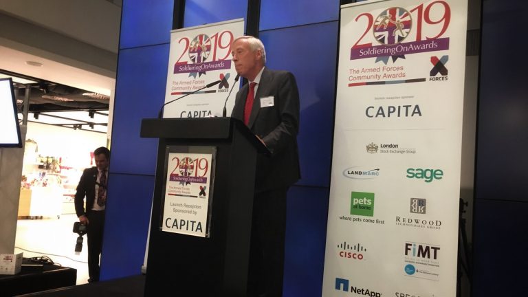 An older man stands on a stage with a microphone, speaking at the Soldiering On Awards event about resettlement programs. Posters with logos are visible in the background, celebrating the commendable journey of those transitioning from Army life.