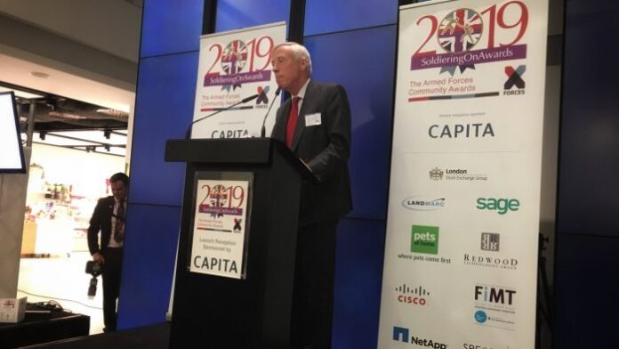 An older man stands on a stage with a microphone, speaking at the Soldiering On Awards event about resettlement programs. Posters with logos are visible in the background, celebrating the commendable journey of those transitioning from Army life.