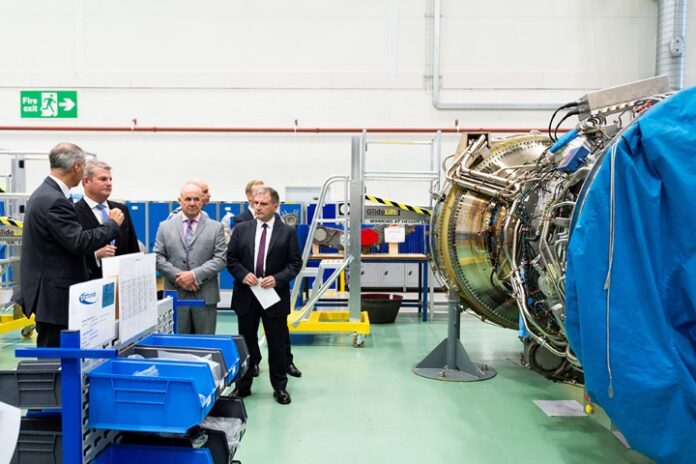 A group of men in suits, like modern-day pathfinders, examine a large industrial machine in a workshop setting.