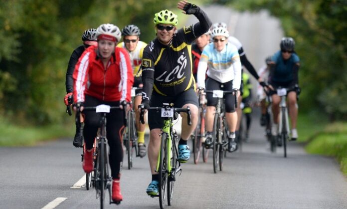 Cyclists riding on a tree-lined road, with one veteran cyclist raising a hand in greeting or celebration.