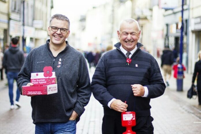 Two men on a bustling street, linked by their pasts in the Royal Navy, collect donations for the Scottish Poppy Appeal. They stand proudly with donation boxes and poppies, embodying the spirit of service to support veterans.