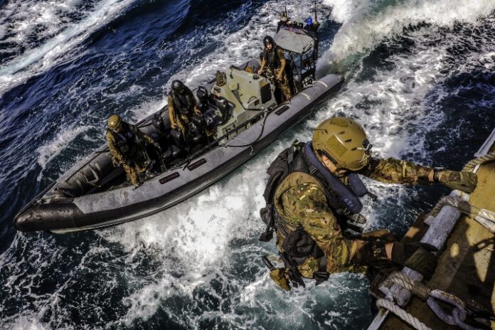 Veteran military personnel in tactical gear, displaying the expertise of the Royal Navy, approach and climb onto a ship from a small inflatable boat on a turbulent sea.