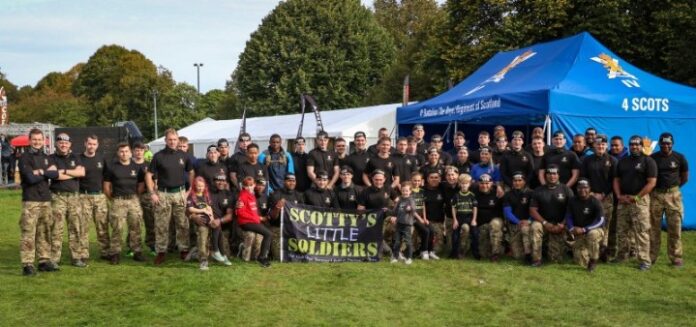 A group of people in camouflage pants and black shirts, resembling a military unit, stand in front of tents on a grassy area, holding a banner that reads 