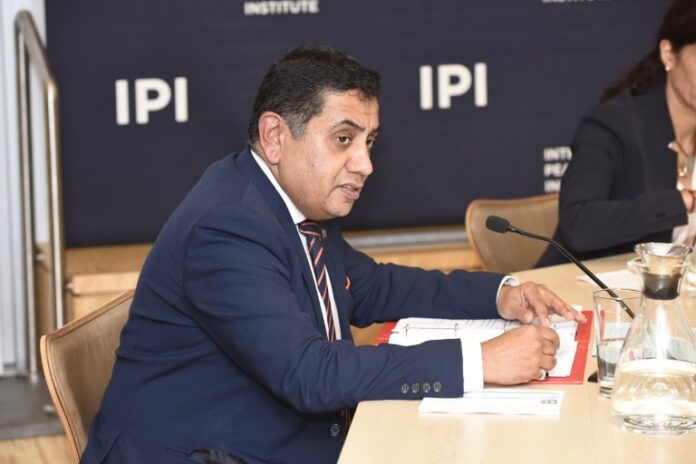 A person in a suit speaks into a microphone at a conference table, with 
