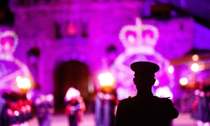 Silhouette of a guard in a hat, reminiscent of an Army pathfinder, facing a brightly lit building adorned with large crown decorations and fellow guards in the background.