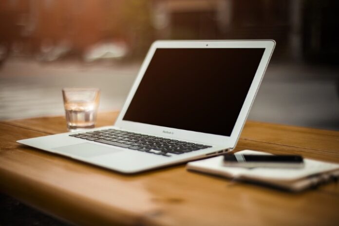 A laptop rests on a wooden table alongside a glass of water, a smartphone, and a notebook—the essentials for a digital pathfinder navigating the modern information battlefield.