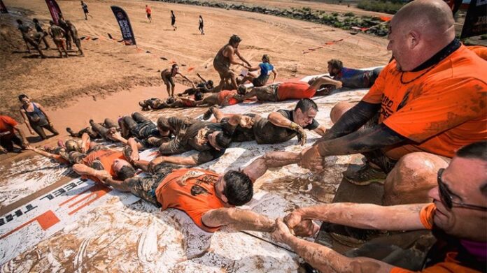Participants in a mud run pull each other up a slippery slope, reminiscent of an Army training exercise, during a team event in an outdoor setting.