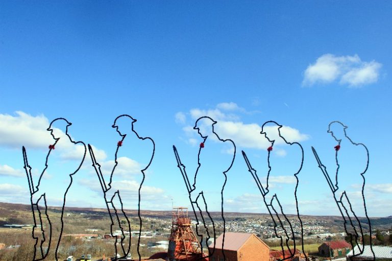 Silhouettes of soldiers with rifles, adorned with red poppies, stand like pathfinders against a scenic view of a hilly landscape under a blue sky.