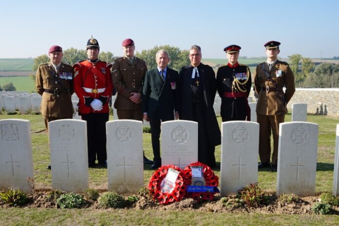 Seven people in uniform stand solemnly behind rededicated soldiers' graves, wreaths placed in front, in a cemetery on a clear day.