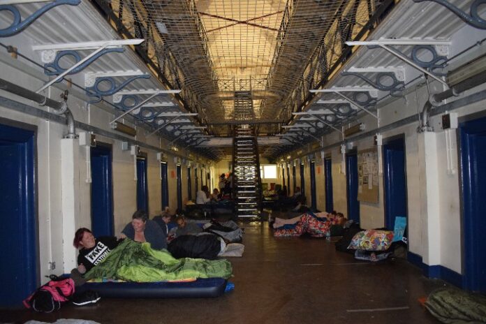 People in sleeping bags and on air mattresses line a dimly lit corridor with metal railings and multiple doors on each side, participating in the BIG Sleep event to raise awareness and funds, their dedication driving success behind every effort.