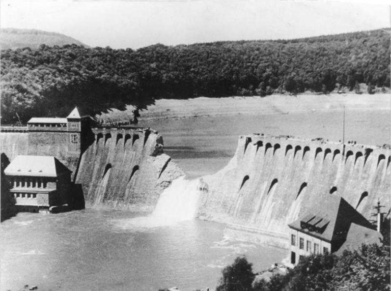 A dam with a large breach in its structure, reminiscent of the daring Dambusters' legacy, releases water into the river below, surrounded by forested hills.