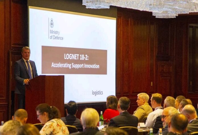 A speaker at a podium addresses an audience in a conference room. The slide on the screen reads 