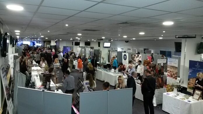 A busy indoor trade show with various booths and people interacting, set in a large, well-lit room; it's an ideal setting for a brilliant networker to showcase their skills and make lasting connections.