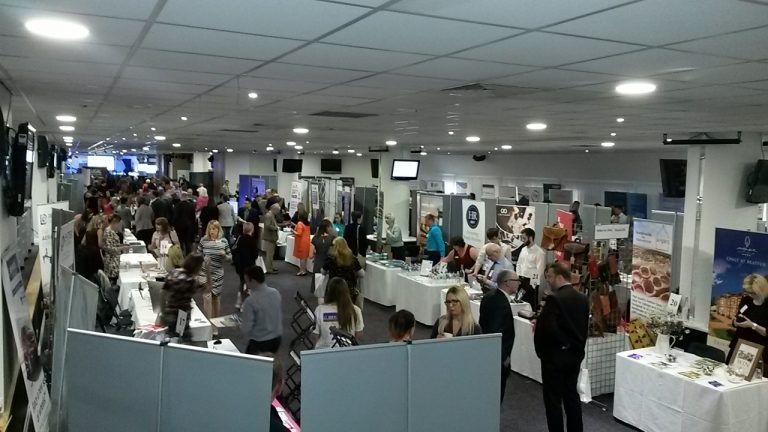 A busy indoor trade show with various booths and people interacting, set in a large, well-lit room; it's an ideal setting for a brilliant networker to showcase their skills and make lasting connections.