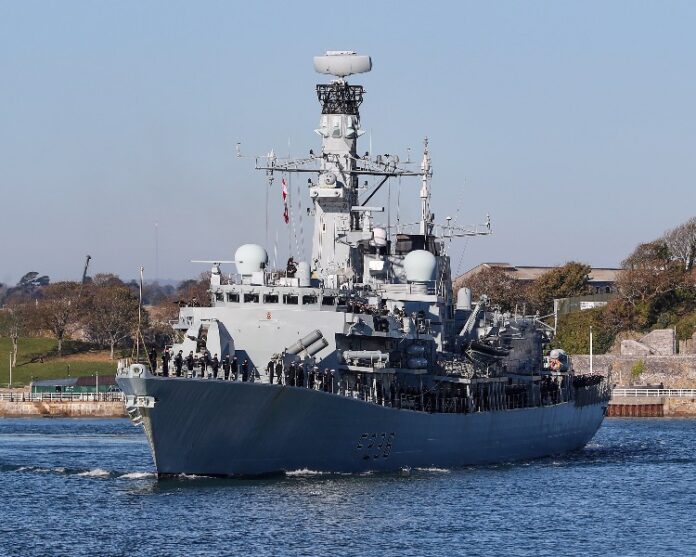 A formidable warship sails on calm water, its crew members clearly visible. The background features a picturesque shoreline with trees and buildings, marking a significant landmark deployment in the Middle East.