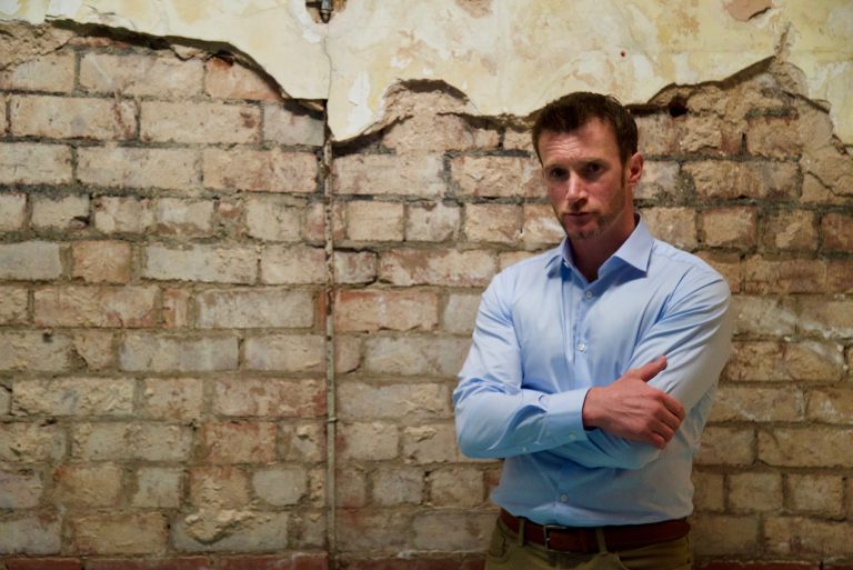 A person in a light blue shirt stands with arms crossed in front of a weathered brick wall, their confidence reminiscent of a pathfinder charting new territories.