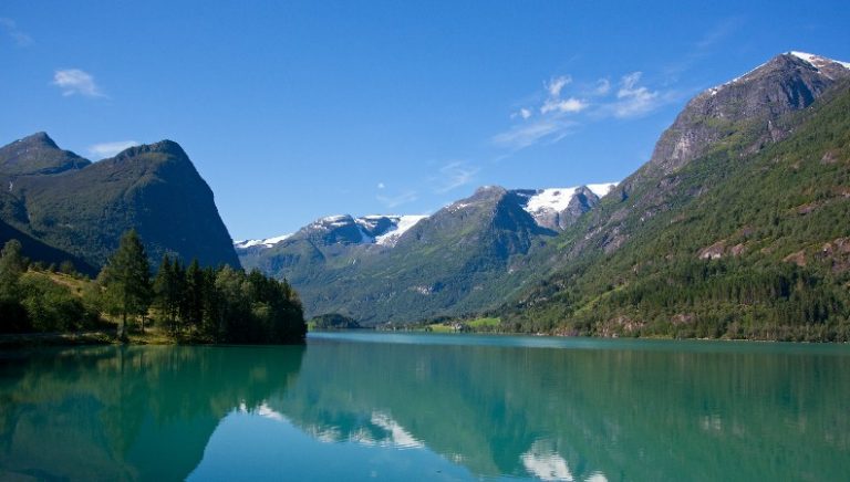A serene lake in Norway with clear blue water reflects surrounding snow-capped mountains and green forests under a clear sky, as UK troops make their road move through the picturesque landscape.
