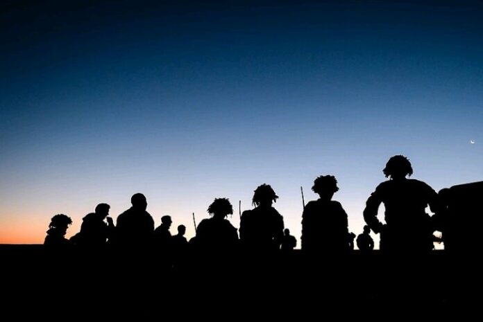 Silhouettes of people, including veterans with gear, stand under a twilight sky against a fading horizon.