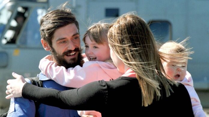 A man smiles while hugging a woman and two children in front of a helicopter, embodying the support network that allows naval families to flourish.