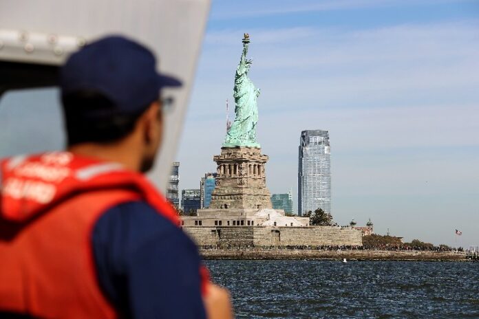 A person in a red life vest looks at the Statue of Liberty across the water, while the HMS Queen Elizabeth majestically passes by, with New York's stunning skyline in the background.