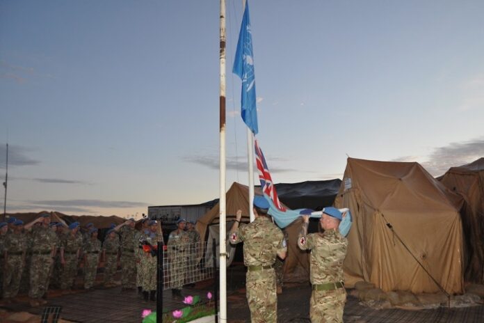 Soldiers in military uniforms, alongside RAF Medics, raise the United Nations and a national flag at a camp with several tents, ready to initiate a transfer operation from a nearby Sudan hospital.