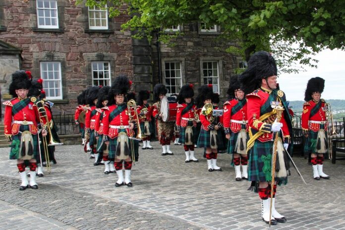 A marching band north of the border, donned in traditional Scottish attire with kilts and feathered hats, expertly performs on a cobblestone path in front of a stone building.