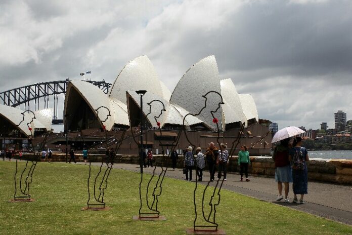 Silhouette sculptures of Tommies adorned with red poppies stand on a lawn near the Sydney Opera House, with pedestrians and the Sydney Harbour Bridge in the background, aiming to raise cash for Invictus Veterans.