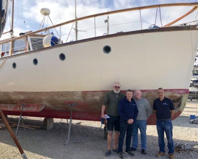 Four men stand in front of a large white boat on dry land, its faded red stripe hinting at its past. With partly cloudy skies above, they discuss their latest project—a boat restoration that is gaining momentum.
