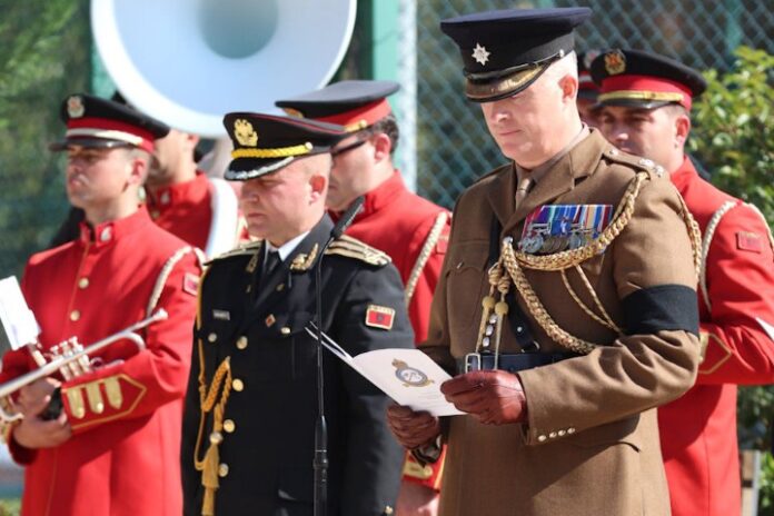 A group of uniformed military personnel stands solemnly, as one holds a document. Several wear red uniforms and play musical instruments, paying tribute to the Halifax Crew laid to rest after serving in WW2.
