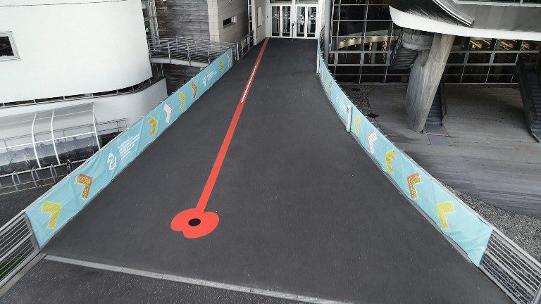 An installation features a pathway with a red line leading to a building entrance, enclosed by blue banners on both sides, reminiscent of the vibrant landscape in Edinburgh, echoing themes often explored by War Poets.