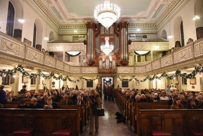 Inside a church, people are seated in pews under chandeliers and festive garlands for the Christmas Carol Concert, with a large organ visible at the front. The warm atmosphere reflects the spirit of giving, reminiscent of Blind Veterans UK's dedication to bringing light into lives.