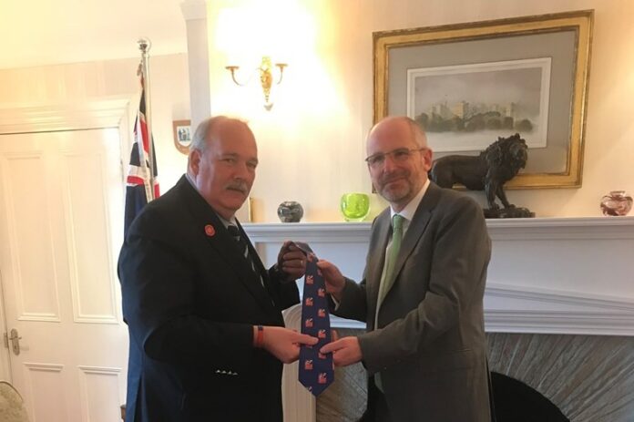 Two men stand in a room, one handing a tie to the other. A flag is in the background, and a lion sculpture is on the mantel. After 36 years, a blind veteran returns from the Falklands to this familiar setting filled with symbols of courage and resilience.