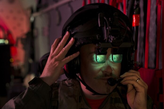 In a dimly lit aircraft, a person wearing night vision goggles and a helmet speaks into a headset, fully immersed in training within the Chinook simulators.