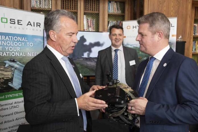 Two men in suits examine a piece of military equipment, essential for the modern battlefield, while a third man watches. Banners in the background display company information, highlighting their role in leading the fight.